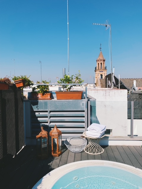 A rooftop terrace with potted plants, a small pool, two lanterns, a chair with towels, and a church tower visible in the background under a clear blue sky—capturing the relaxed elegance often found in boutique hotels in Seville.