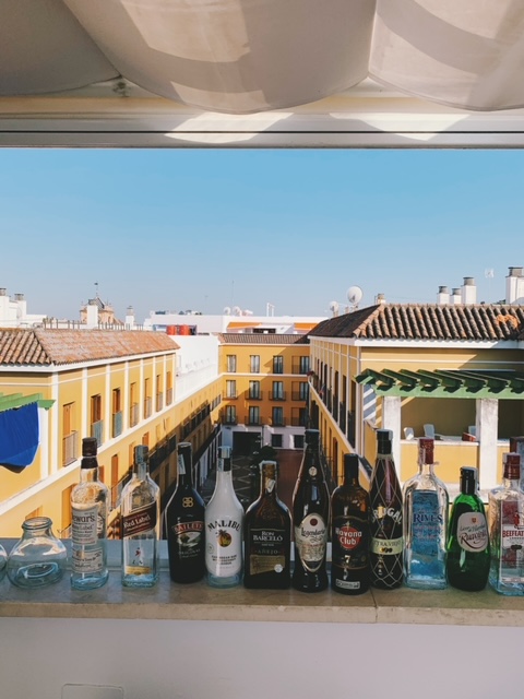 A row of liquor bottles lines a balcony ledge, overlooking a courtyard with yellow buildings, tile roofs, and a blue sky in the background—an inviting scene that captures the vibrant atmosphere often found at boutique hotels in Seville.